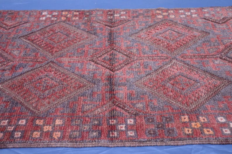 Op de afbeelding: Een rechthoekig vloerkleed met een herhalend patroon van grote ruiten in tinten dieprood en gedempt blauw. Het ontwerp bevat kleinere geometrische motieven en een rand met een raster van rode, blauwe en cr&egrave;mekleurige vierkantjes.