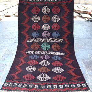 May include: A dark brown rectangular rug with a geometric pattern. The rug features repeating diamond shapes in various colors, including red, white, and brown. The edges have a decorative border with fringe.