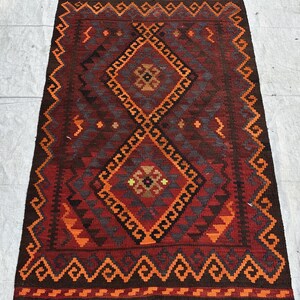 May include: A rectangular, hand-woven rug with a geometric design. The rug features a central diamond pattern in shades of red, orange, and brown, framed by a border of repeating geometric shapes. The rug has a fringed edge.