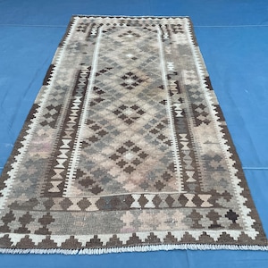 Pode incluir: Tapete retangular com um padrão geométrico de losangos em tons de castanho, bege e cinza. O tapete tem uma borda com formas geométricas repetidas e franjas nas extremidades. Um tapete em tons neutros.