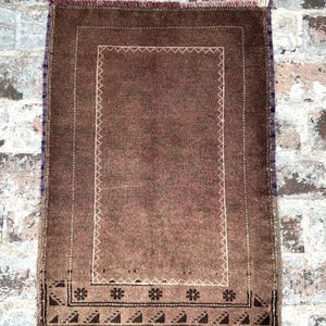 May include: A rectangular brown rug with a central field and a decorative border. The rug features a central rectangular design with a white stitched border. The outer border has floral and geometric patterns.