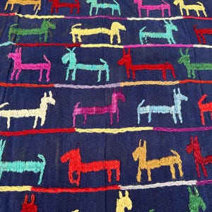 May include: Close-up of a dark blue fabric with embroidered donkey figures in various colors. The donkeys are arranged in rows, with horizontal lines of embroidery in different colors separating the rows. The embroidery is detailed and colorful.