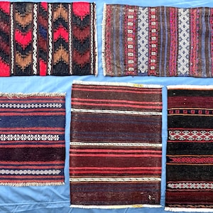 May include: A collection of small, rectangular woven rugs with intricate patterns. The rugs feature a variety of colors, including red, black, brown, and blue, with geometric and striped designs. Some have fringed edges.