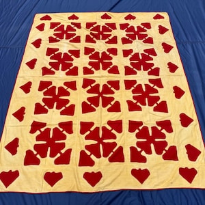 May include: A rectangular quilt featuring a cream-coloured base with a repeating pattern of deep red, four-petaled floral or heart-shaped motifs arranged in a grid. The edges of the quilt are finished with a thin, solid red fabric border.