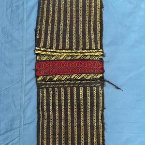 Pode incluir: Têxtil retangular tecido com fundo castanho escuro e riscas verticais amarelas. O centro apresenta um padrão geométrico vermelho. As bordas têm franjas coloridas em laranja, amarelo e verde. O têxtil é provavelmente feito à mão.