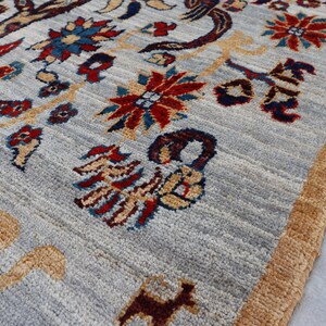 May include: A hand-knotted area rug featuring a light gray background with a repeating pattern of stylized floral motifs in red, blue, and yellow, alongside animal figures in tan and dark brown, finished with a thick, textured tan border.
