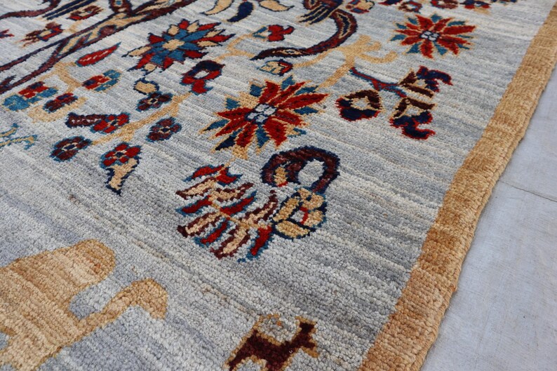May include: A hand-knotted area rug featuring a light gray background with a repeating pattern of stylized floral motifs in red, blue, and yellow, alongside animal figures in tan and dark brown, finished with a thick, textured tan border.