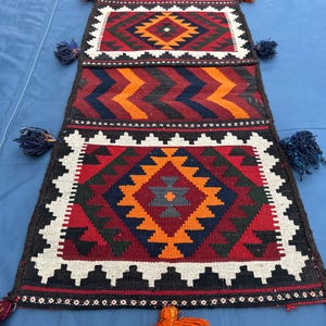 Pode incluir: Tapete tecido à mão com um design geométrico. O tapete apresenta um padrão central de diamante em laranja, vermelho e azul, emoldurado por uma borda branca e preta. Tassels adornam as extremidades. Têxtil tradicional.