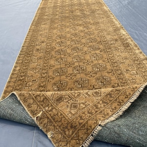 May include: A long, rectangular runner rug featuring a tan and brown color palette with a repeating geometric pattern of octagonal motifs. The rug has a visible fringe at the end and is folded over to reveal a dark, textured backing material.