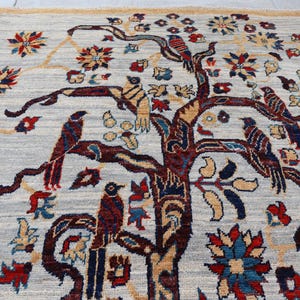 May include: A rectangular area rug featuring a light gray background with a central dark brown tree motif. The tree branches are adorned with various birds in shades of red, blue, and brown, interspersed with colorful floral patterns in red and blue.