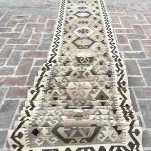Puede incluir: Una alfombra de pasillo larga y estrecha con un patrón geométrico repetitivo en tonos beige, marrón, crema y gris carbón. La alfombra tiene un borde decorativo con un motivo en zigzag y está rematada con flecos cortos y rectos en el borde.