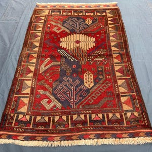 May include: Hand-knotted oriental rug with a central design in red, navy blue, and beige. The rug features geometric patterns and a fringed edge. The overall design is symmetrical and traditional, suitable for home decor.