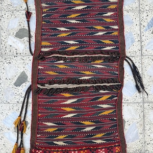 May include: A rectangular, woven textile with a geometric pattern in red, black, yellow, and white. The textile has brown trim and tassels. The design features repeating diamond and chevron shapes. The textile is likely a decorative item or a bag.
