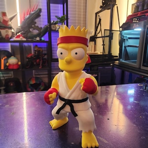 May include: A yellow figurine of Bart Simpson in a karate outfit. The figure wears a red headband and gloves, a white gi with a black belt, and yellow feet. The figure is posed in a fighting stance.