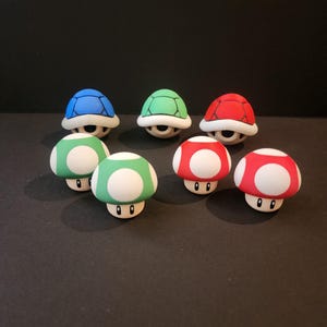 Puede incluir: Una colección de figuras coloridas con temática de Super Mario. La imagen presenta seis figuras de champiñones en rojo y verde con puntos blancos, y tres figuras de caparazones de tortuga en azul, verde y rojo. Todas las figuras tienen una base blanca.