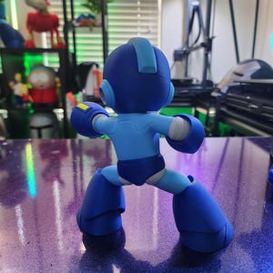 Puede incluir: Una figura de robot azul y amarillo con un casco y un cuerpo azul. El robot est&aacute; de pie con las piernas separadas y los brazos levantados.