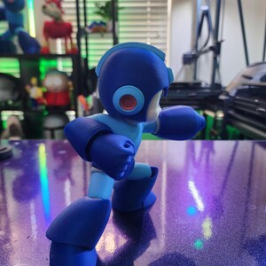Puede incluir: Una figura de acci&oacute;n de pl&aacute;stico azul y azul claro de un robot con una cabeza grande y un solo ojo rojo. El robot est&aacute; en posici&oacute;n de combate con los brazos levantados.