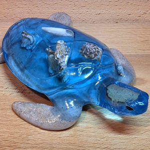 Peut inclure: Une figurine de tortue en résine bleue avec des coquillages incrustés. La carapace de la tortue est d'un bleu translucide, et les nageoires sont gris moucheté. L'objet est décoratif, probablement pour la décoration intérieure.