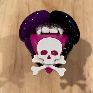 May include: A black and purple lips phone charm with a pink tongue and a white skull and crossbones design.