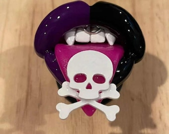 May include: A black and purple lips phone charm with a pink tongue and a white skull and crossbones design.