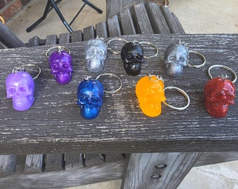 May include: A collection of skull-shaped keychains in various colors, including purple, silver, black, blue, orange, and red. Each keychain has a silver ring. The keychains are displayed on a weathered wooden surface.