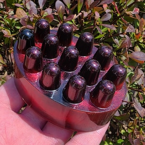 May include: A red and black scalp massager with a glittery finish. The massager has a series of rounded, dark red nodes. One node is black. The massager is held in a hand against a background of green and red foliage.