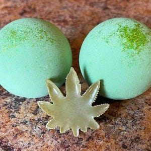 Puede incluir: Dos bombas de baño verdes con un diseño de hoja verde. Las bombas de baño son redondas y tienen una textura moteada.