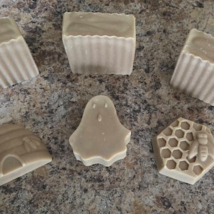 May include: A selection of handmade soaps in different shapes: a ghost, a beehive, a honeycomb with a bee, and rectangular bars with a scalloped edge. The soaps are a light beige colour, arranged on a speckled surface.