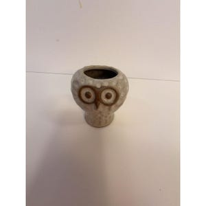 May include: A small, round ceramic vessel shaped like an owl. The piece features a speckled off-white glaze with a textured surface. The owl's face is defined by two prominent, circular brown eyes and a small, pointed beak centered on the front.