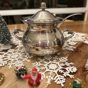 May include: A silver-colored, ornate sugar bowl with a lid and a small spoon. The bowl has two handles and rests on three small feet. The design includes decorative patterns. The bowl is part of a holiday-themed display with snowflake decorations and a Santa Claus figurine.