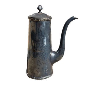 May include: A silver-coloured metal coffee pot with a long, curved spout and a lid. The pot has a cylindrical body with decorative trim around the base and lid. The word "Duchesse" is engraved on the side.