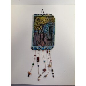 May include: A rectangular wall hanging with a painted scene of a figure under a tree, with a tall, dark object. The artwork is in shades of yellow, blue, purple, and black. Decorative beads hang from the bottom.