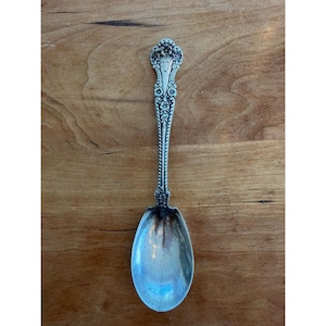 May include: A silver spoon with an ornate floral design on the handle and a long, slender stem. The spoon's bowl is oval-shaped and reflects the light. The spoon is likely vintage or antique, and is set against a wooden background.