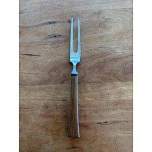 May include: A vintage carving fork with a two-pronged stainless steel head and a wooden handle. The handle is a medium brown color and tapers slightly towards the end. The fork is designed for serving meat.