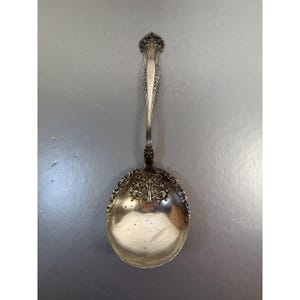 May include: A silver serving spoon with ornate detailing. The spoon features a decorative handle and a bowl-shaped head with intricate floral and scrollwork patterns. The spoon is likely made of sterling silver and is designed for serving food.