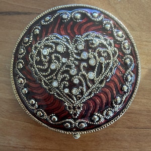 May include: A round, decorative compact mirror with a red and gold design. The compact features a heart-shaped filigree design with embedded rhinestones. The border is gold with small, decorative accents.
