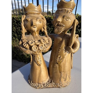 May include: A ceramic sculpture depicting a royal couple. The figures are tan-colored, with the woman holding a bouquet of roses and wearing a crown. The man has a beard, a crown, and holds a staff. The base is textured.