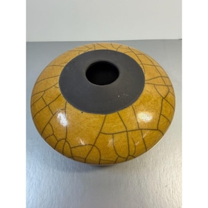 May include: A round ceramic vase with a yellow-brown crackle glaze pattern. The vase has a dark gray, circular opening at the top. The vase is a decorative item.