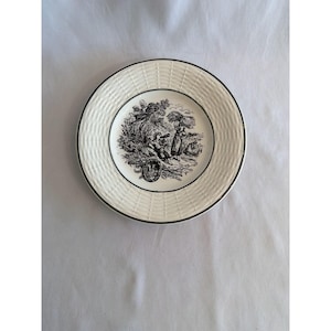 May include: A cream-coloured ceramic plate featuring a black and white rural scene. The plate has a woven texture around the edge and a black trim. The scene shows figures in a field.