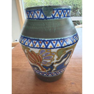 May include: A ceramic vase with a wide base and a narrow neck. The vase features a green band around the middle and decorative patterns in blue, orange, and purple. The top rim has a chip. The vase is on a wooden surface.
