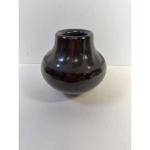 Könnte beinhalten: Eine glänzende, dunkelbraune Keramikvase mit rundem Körper und ausgestelltem, zylindrischem Hals. Die Vase hat eine glatte, reflektierende Oberfläche, und die Öffnung ist dunkel gefärbt. Die Vase dient wahrscheinlich dekorativen Zwecken.