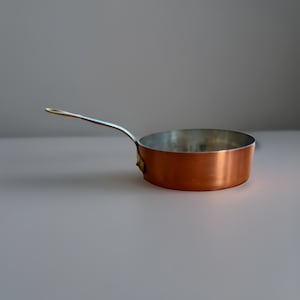 May include: A small copper saucepan with a silver interior and a long, silver handle. The pan has a rounded shape and a shiny, reflective surface. The handle is attached to the side of the pan.