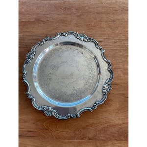 May include: A silver serving tray with a decorative, scalloped border. The tray features a circular central area with a delicate, embossed floral design. It sits on a wooden surface, highlighting its reflective nature and vintage style.