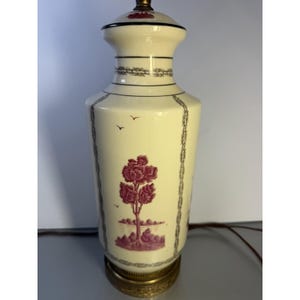 May include: Cream ceramic table lamp with a decorative design. The lamp features a maroon tree and landscape scene, with black and grey accents. The base is a gold-coloured metal.