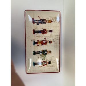 May include: A rectangular ceramic serving tray with a cream-coloured background featuring a vintage script and postmark pattern. The tray displays five colourful nutcracker figures in various uniforms, bordered by a thin, dark red rim.