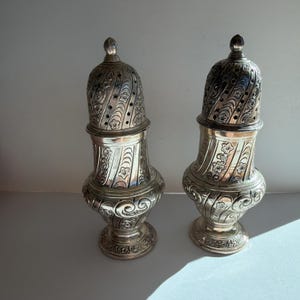 May include: Two ornate silver salt and pepper shakers. Each shaker has a bulbous base, a detailed floral and scroll design, and a perforated, domed top. The shakers are displayed on a white surface, with sunlight casting shadows.