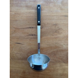 Könnte beinhalten: Eine Edelstahl-Schöpfkelle mit schwarzem Griff. Die Schöpfkelle hat eine runde, tiefe Schüssel und einen langen, schlanken Griff. Der Griff ist mit einem Metallstreifen an der Schüssel befestigt. Zum Servieren von Suppen, Eintöpfen und Saucen.