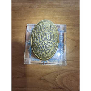 May include: An antique brass-coloured oval plaque with the words "PUBLIC SCHOOL CITY OF NEW YORK" in an ornate font. The plaque is set on a clear, rectangular block with blue floral designs.