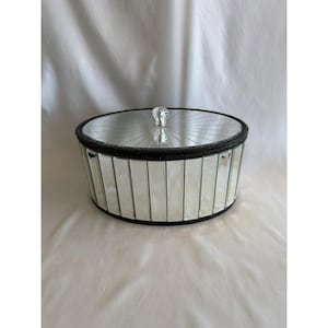 Puede incluir: Joyero ovalado con espejo, borde negro y pomo de cristal en la tapa. La caja está hecha de paneles de vidrio espejado. La tapa también es espejada.