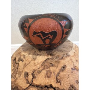 Op de afbeelding: Een handgemaakte, donkerbruine keramische kom met een roodbruin middenstuk met een zwart dierfiguur en geometrische patronen. De kom staat op een natuurlijk houten oppervlak, wat de unieke details benadrukt.